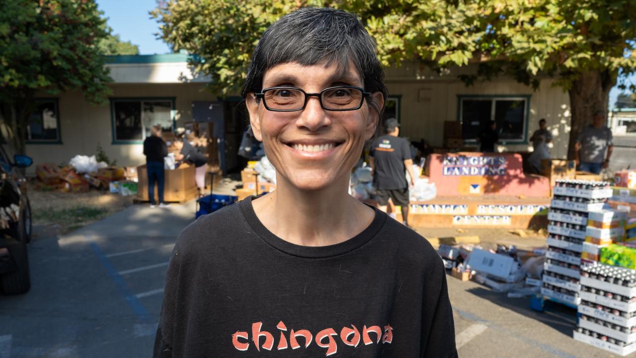 A person with short hair and glasses smiles while standing outdoors. They are wearing a black shirt with the word "chingona" printed on the front. In the background, people are working near tables with boxes and bags, with a building and trees visible.