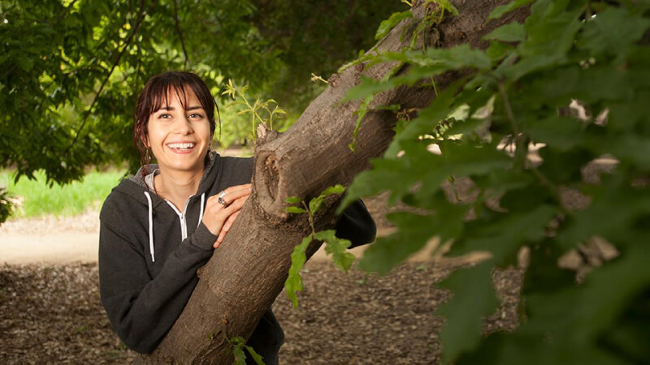Woman smiling widely. She is wearing sweater and is standing outside behind a tree.