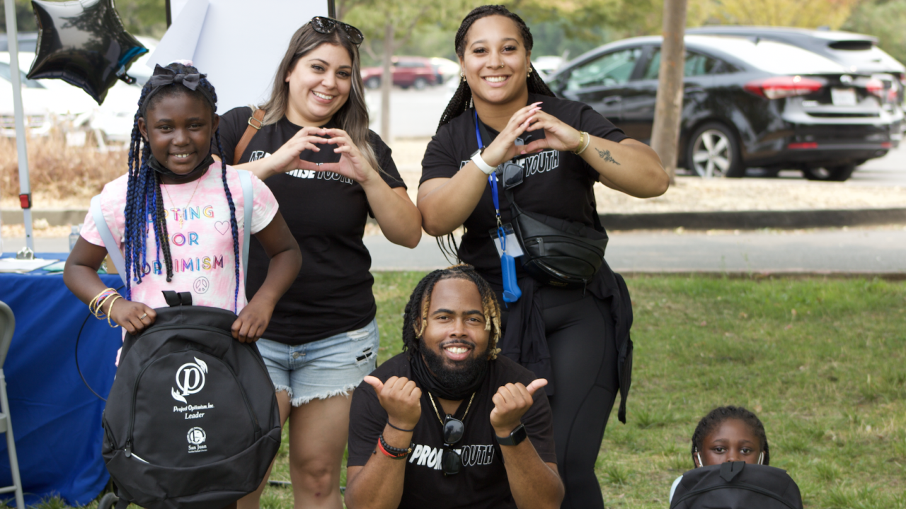 A group of five people pose outdoors. Two adults standing in the back form heart shapes with their hands, wearing black "Promise Youth" shirts. A young girl in the front left smiles, wearing a pink shirt that says "Fighting for Optimism" and holding a black backpack. In the center, a man crouches, smiling and giving two thumbs up, also wearing a "Promise Youth" shirt. Another child sits on the ground to the right, partially hidden behind a backpack. Cars and trees are visible in the background.