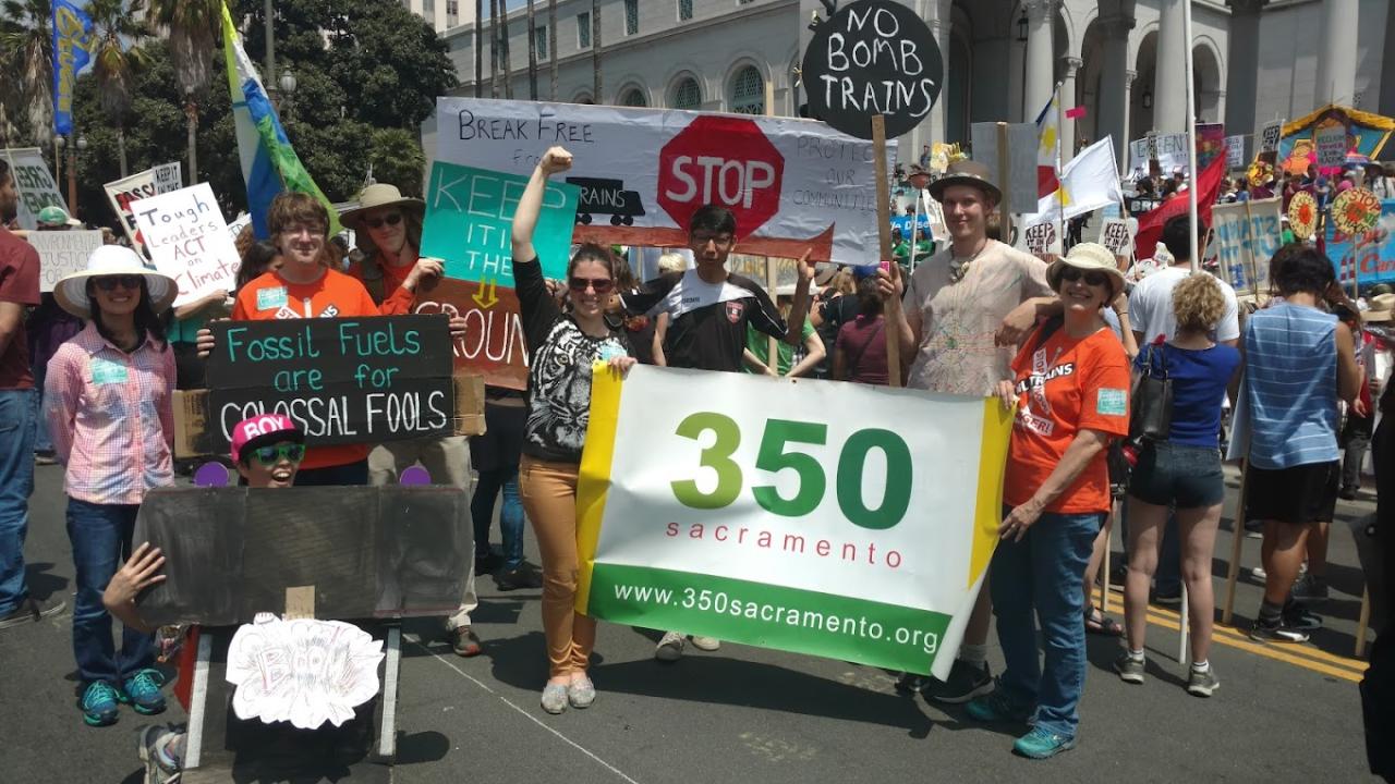 Crowd protest in front of a building while holding various signs that say "No Bomb Trains" and "Fossil Fuels are for Colossal Fools" as well as a banner that says "350 Sacramento"