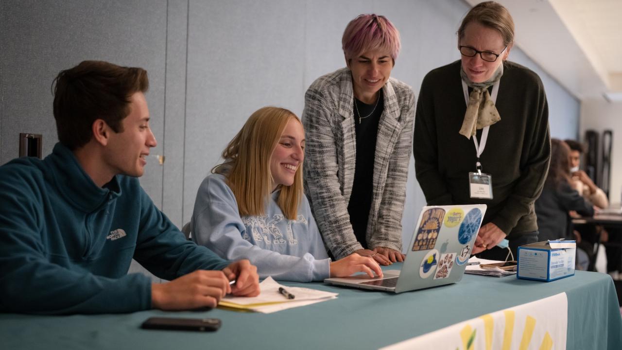 Four people gather around a table with a laptop. One person sits on the left, smiling, with a pen and notebook in front of them. Another person, seated in the middle, types on the laptop. Two people stand behind the laptop, looking at the screen. The table is covered with a teal cloth and a banner featuring a sun and plant graphic. The background shows other people sitting at tables.