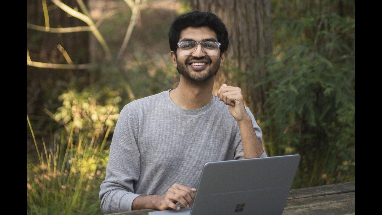 uc davis computer science student wearing glasses sits in front of a tree at a bench with his computer on the table