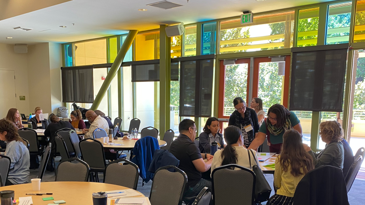 A photo showing the community engagement summit at UC Davis. People are sitting at round tables and engaged in conversation. A few people are standing along the edge of the room. Colored windows in yellow, orange and blue are overhead.