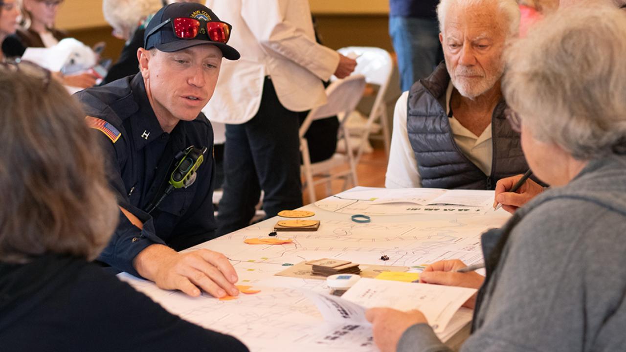 A group of adults gathered around a table play a board game that looks like a map of the Tomales community