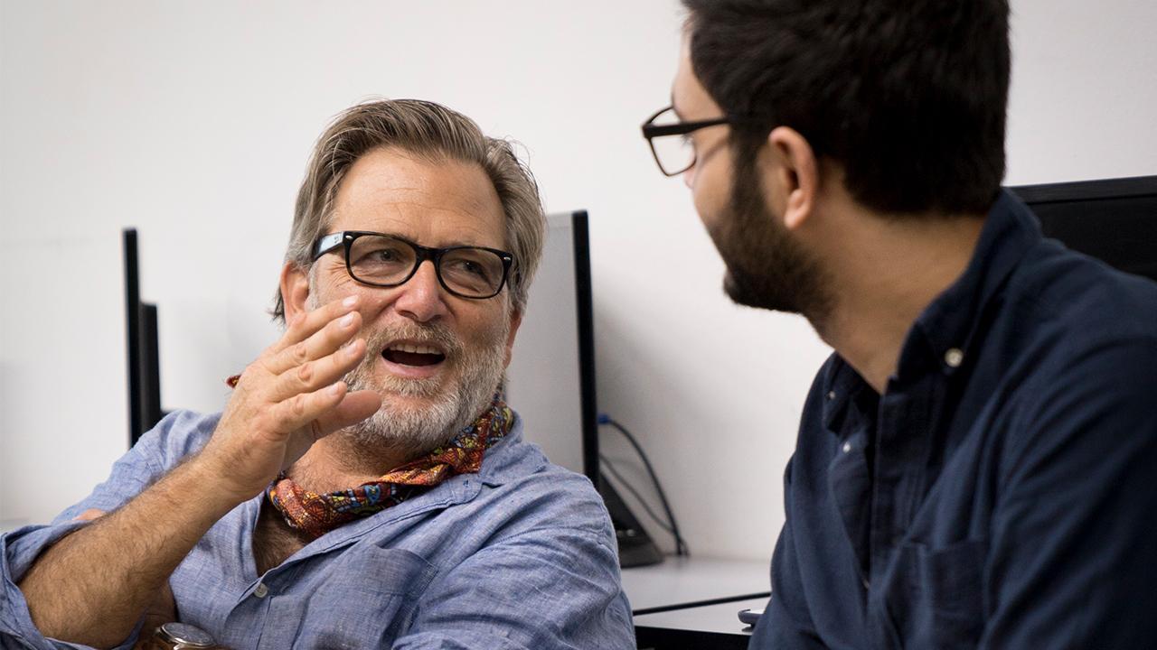 a man in a blue shirt and glasses with his hand raised talking to a younger man