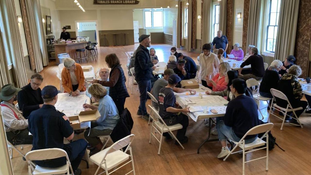 Five groups of individuals gather around five separate table playing a board game indoors at the Tomales Branch Library