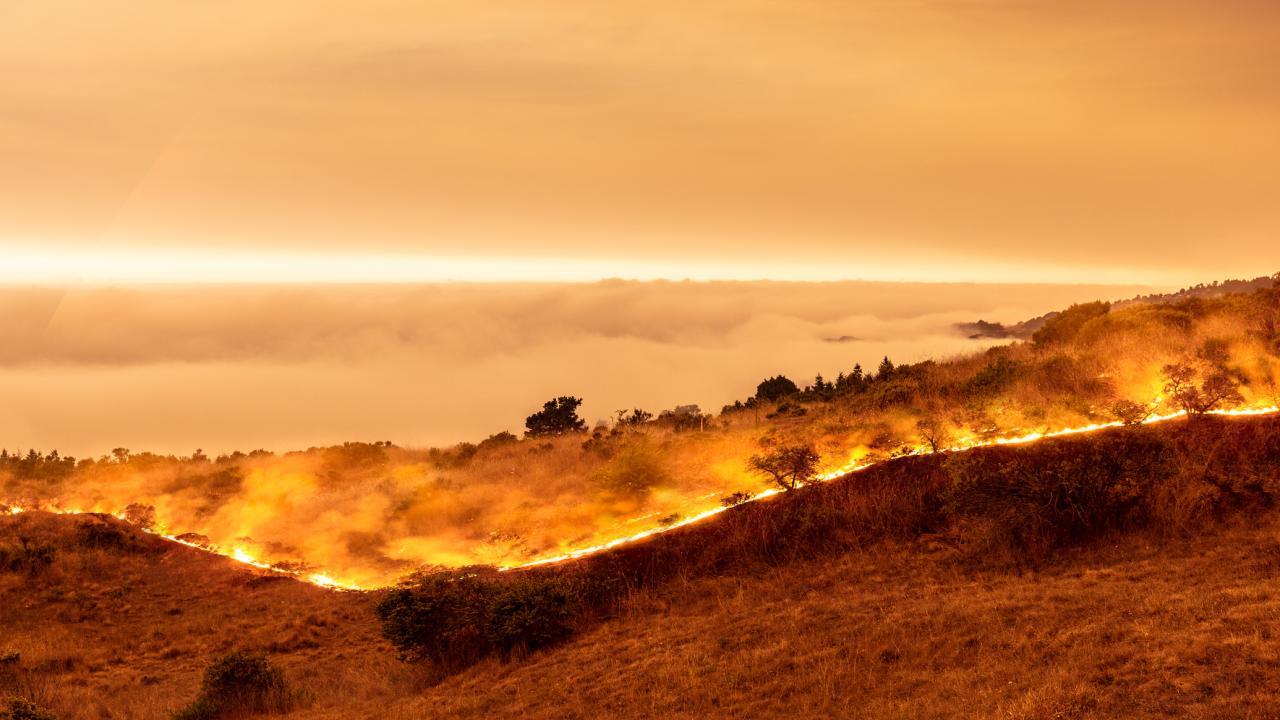 Orange smoky sky with marine layer and line of wildfire in California coastal hills