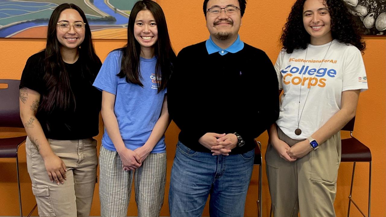 Four individuals stand side by side smiling for a close up group photo in front of a wall that has two artwork peeking on both the left and right side