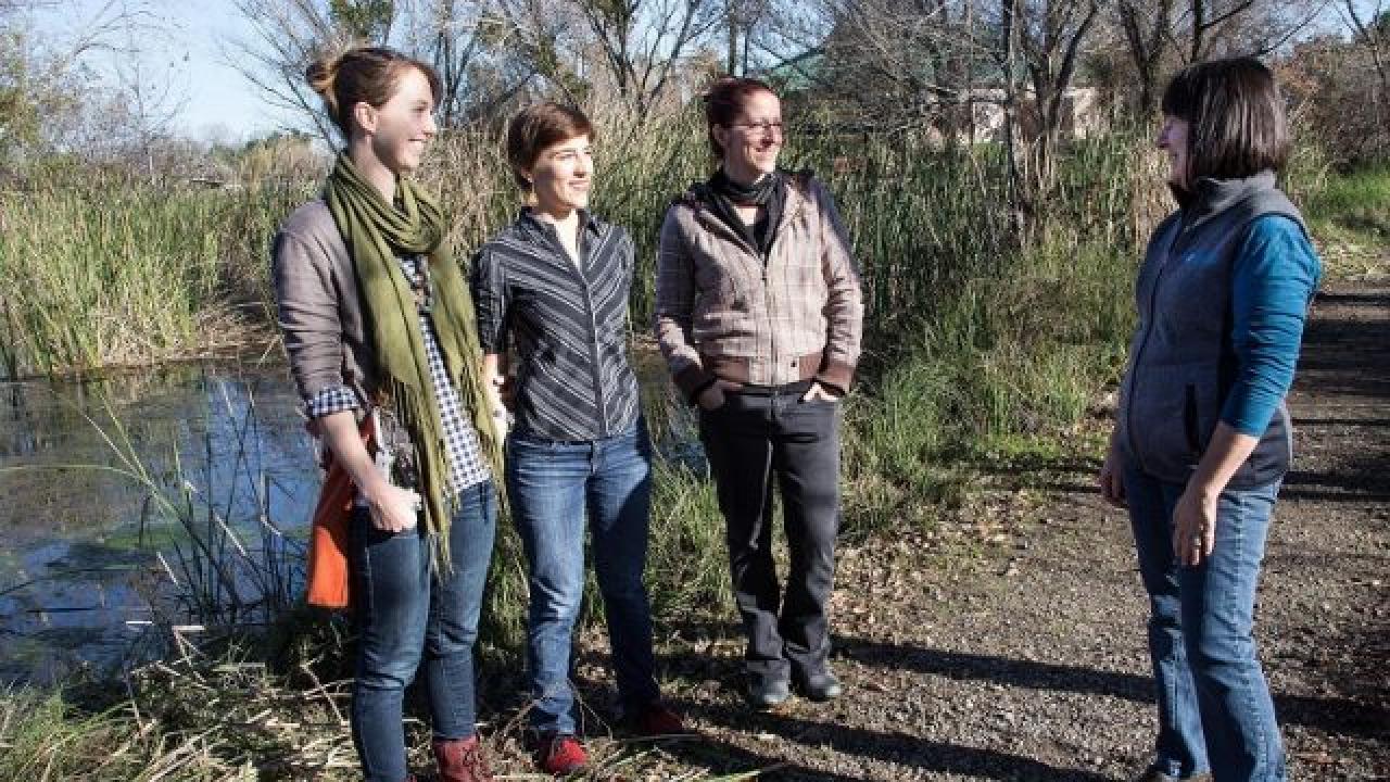Three individuals smile together while facing towards a woman who is also smiling. The group appears to be in discussion outside on a trail.