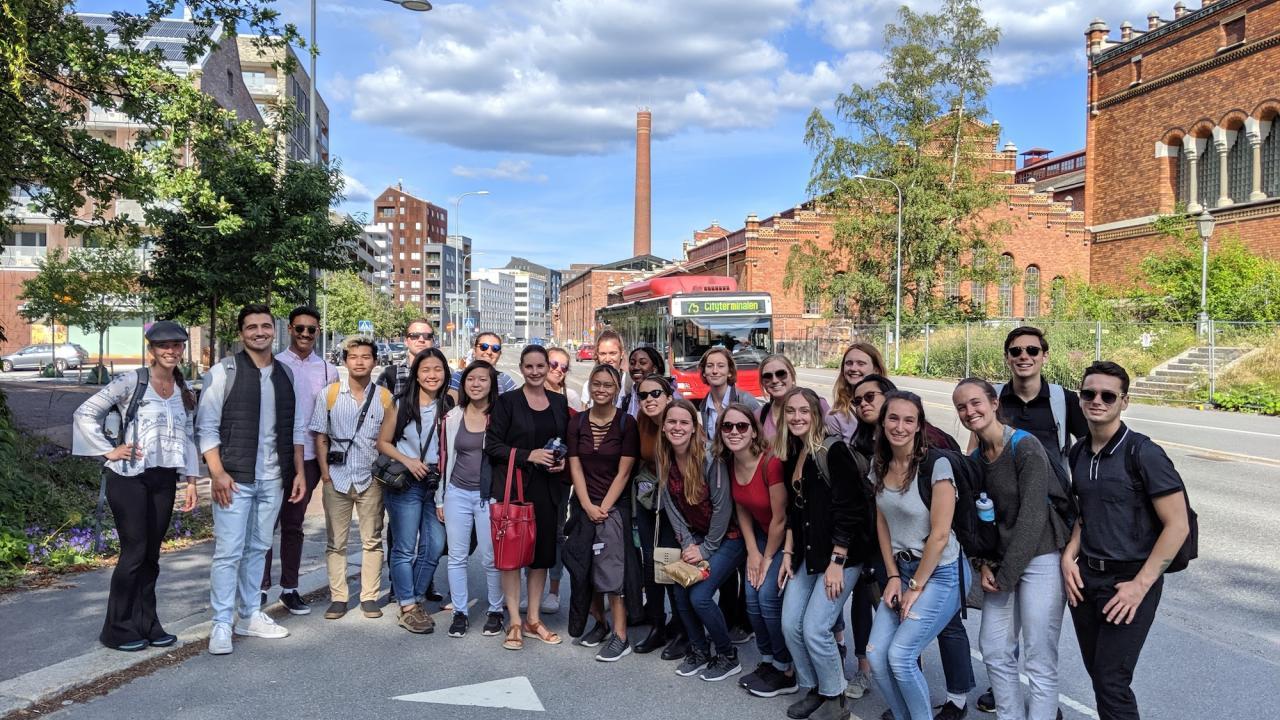 a large group of young people posing for a photo on a street corner with brick buildings and a bus in the background