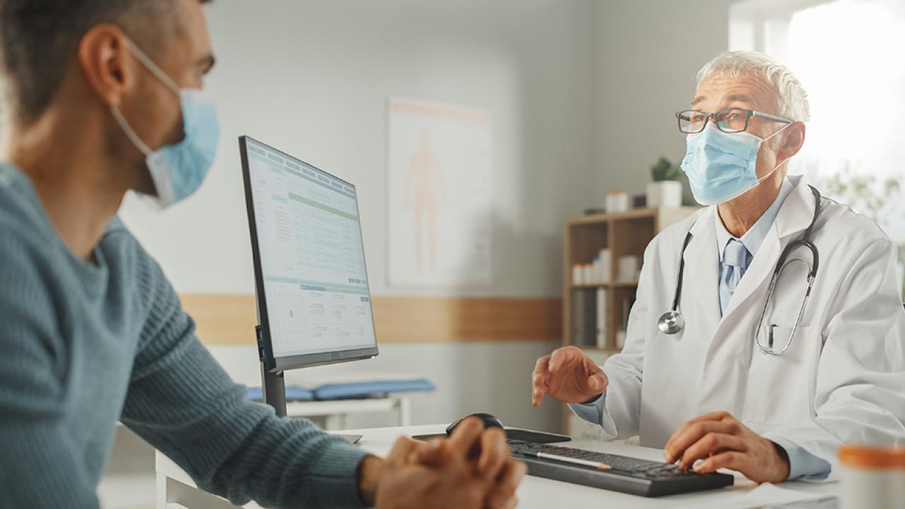 a white haired doctor in a mask wearing glasses talking to a patient in a blue sweater wearing a mask