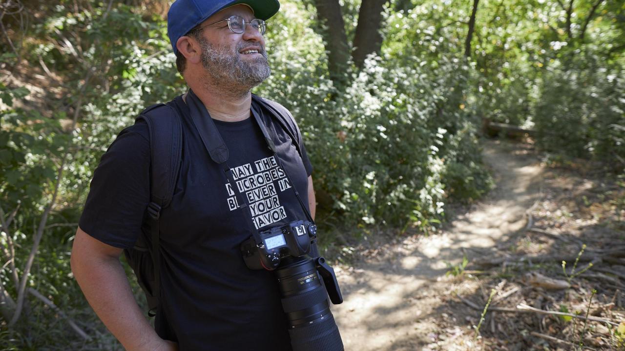 a man in a blue hat and glasses stands against a verdant green backdrop with a camera around his neck