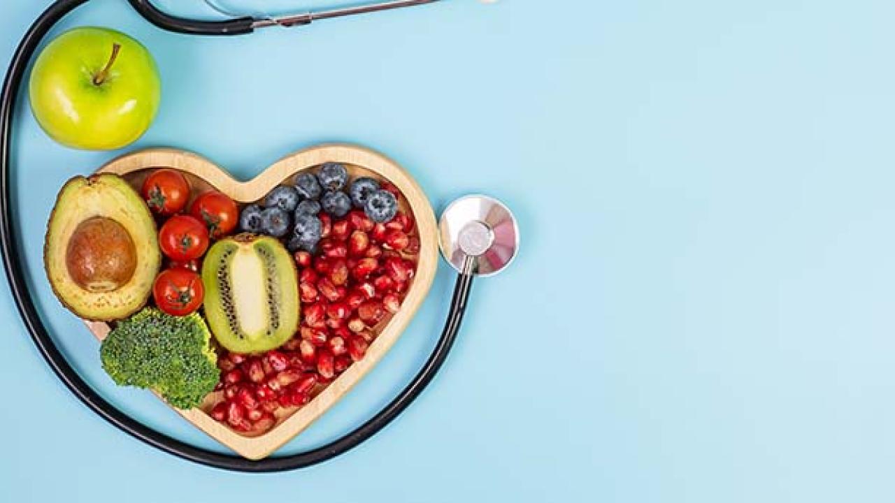 Fruit and vegetables in a wooden heart-shaped box next to a stethoscope and an apple.