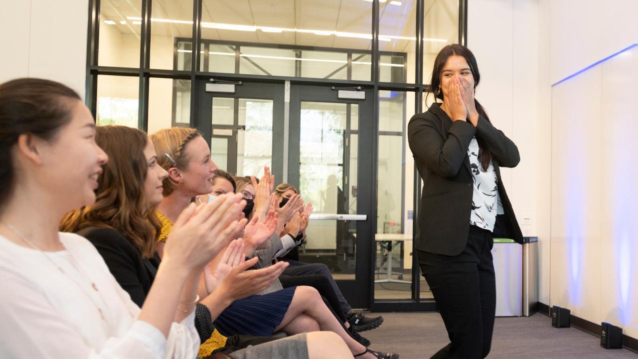 Woman holds hands to mouth, expressing surprise in front of an audience clapping