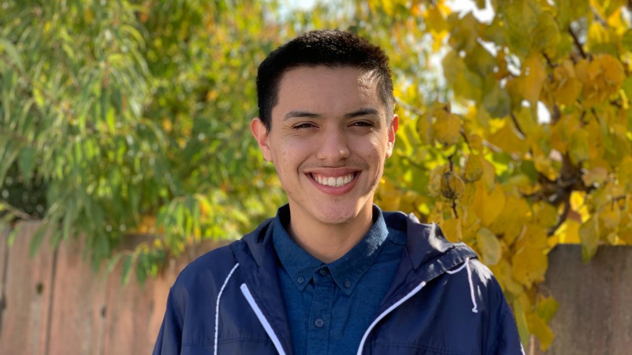 a young latinx man smiling with trees and a fence behind him 