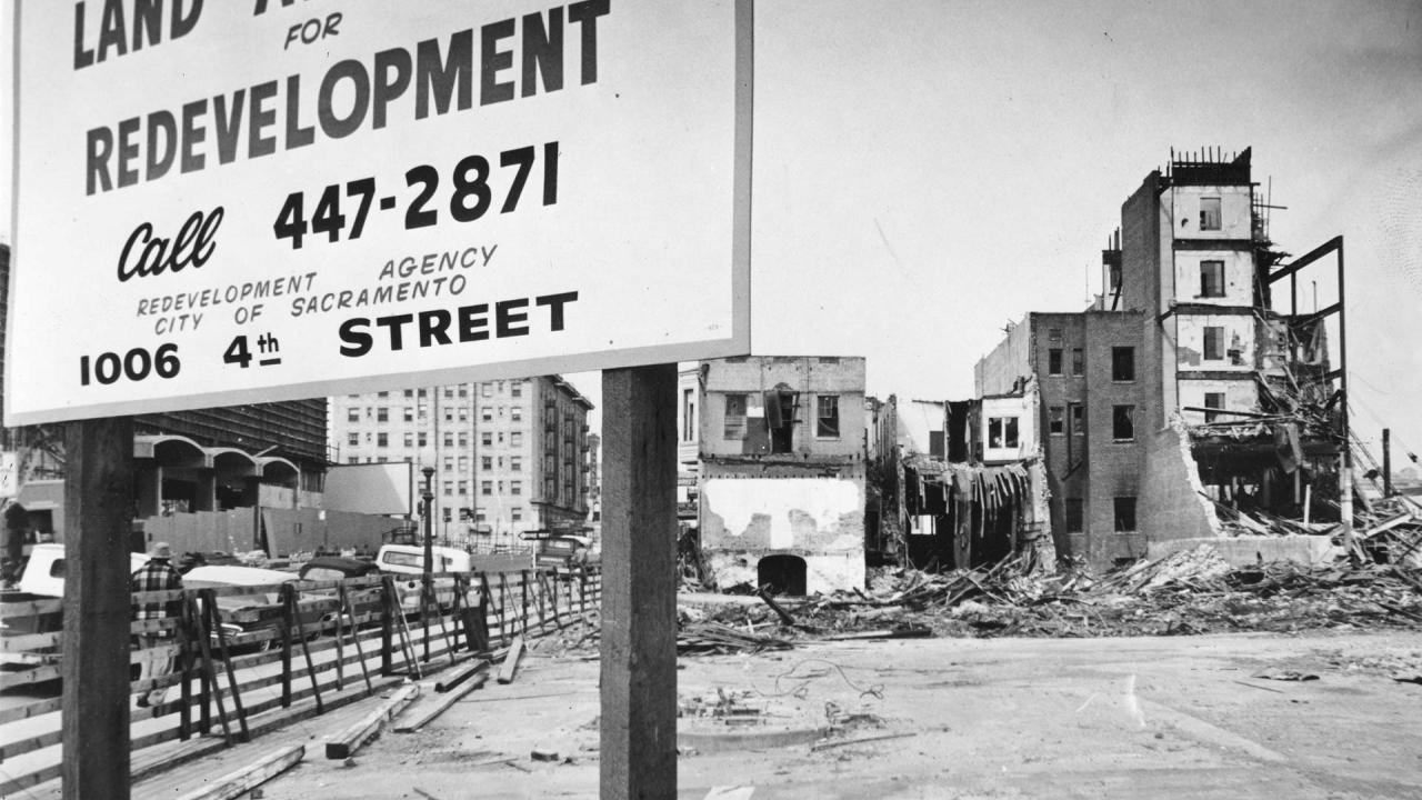 Black and white photo showing a large sign put up in front of a vacant lot. The sign reads "Land Available for Redevelopment" and gives a phone number and address. Buildings toward the back of the lot are undergoing various stages of demolition.