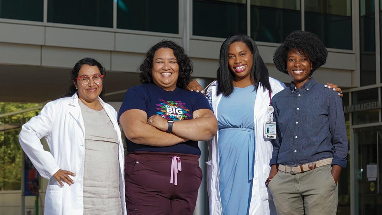 A group of four women stand together in front of a medical office building. Two wear lab coats and one wears scrubs. One has a stethoscope. All are smiling and looking directly at us.