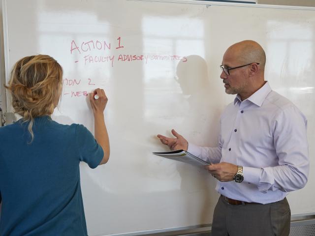 a woman writing on a white board as a man talks to her