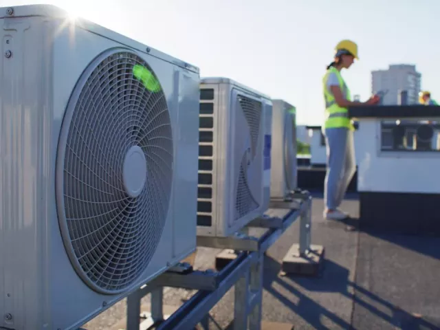 Oblique view of air conditioner units on a rooftop. A technician wearing a high visibility vest and hard hat is in the background. 
