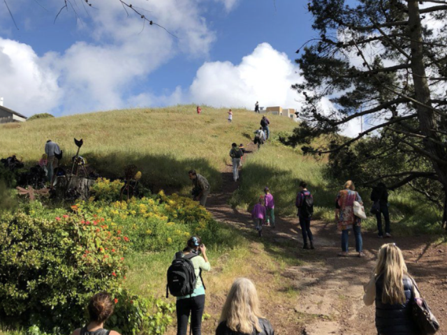 Group of individuals walk on a trail going up a hill. The sky is clear with clouds. 