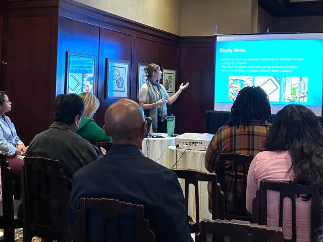 A group of people sits attentively in a conference room, watching a presenter who is standing next to a projection screen. The presenter, wearing a scarf and gesturing with one hand, is discussing a slide titled "Study Aims." The slide includes bullet points and colorful images. Attendees, seated in rows, focus on the presentation, some taking notes. A projector sits on a table in front of the screen, and framed artwork is visible on the wall behind the group.
