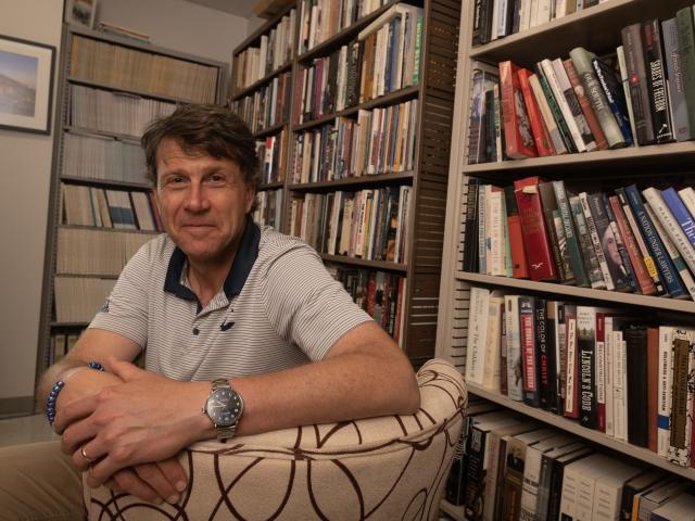 Man in a collared shirt is sitting in a chair. He turns around to smile for a photo while looking directly into the camera. He sits in front of shelves of books.