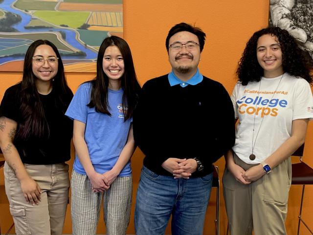 Four individuals stand side by side smiling for a close up group photo in front of a wall that has two artwork peeking on both the left and right side