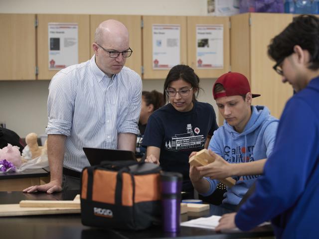  Man is looking at a computer screen from a student’s computer inside of a classroom. Two other students to the side are working on a wood project together. 