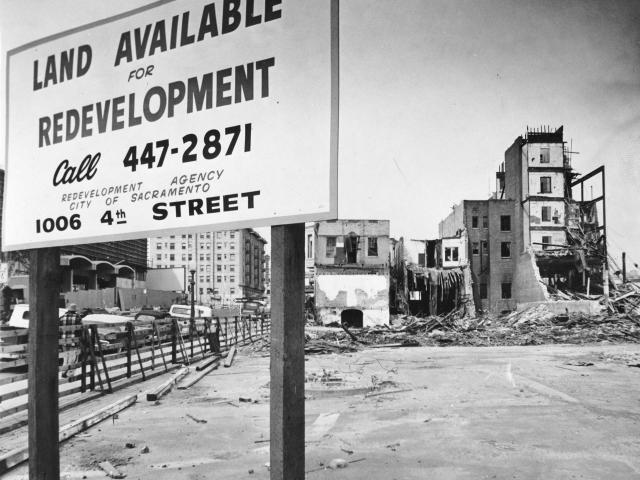 Black and white photo showing a large sign put up in front of a vacant lot. The sign reads &quot;Land Available for Redevelopment&quot; and gives a phone number and address. Buildings toward the back of the lot are undergoing various stages of demolition.