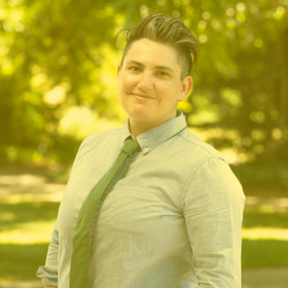 A person wearing a shirt and tie stands outdoors, with short hair styled to one side. The background shows greenery.