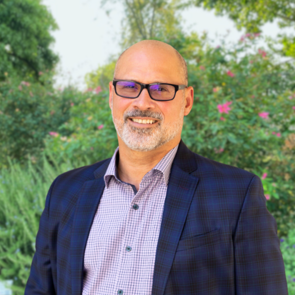 Michael Rios, man wearing tinted glasses and a plaid blazer and checkered dress shirt smiling directly into the camera outside in front of bushes of flowers and trees.