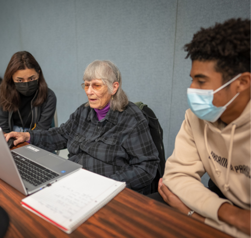 Two student sit side by side with an older adult to teach her how to use the computer.