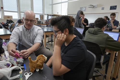 a man with no hair and glasses talking to a student holding stuffed animals