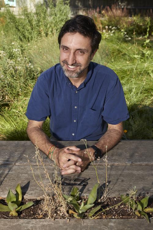 a man in a blue shirt with hands crossed with greenery in the background