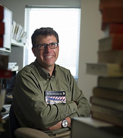 Man wearing glasses and a button down shirt holds books closely to his chest to smile for a photo inside an office with stacks of books