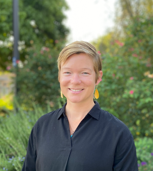 Amy Hart, woman with short hair wearing a collared shirt smiles directly into the camera outside in front of bushes of flowers and trees.