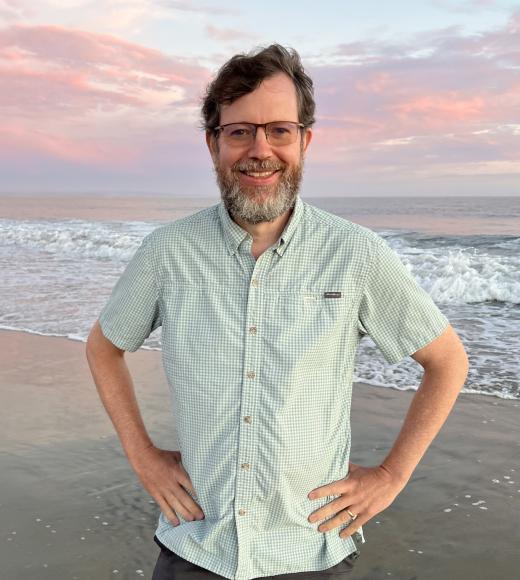 Man smiling and standing at the beach on the sand in front of the sunset ocean with his hands on his hips