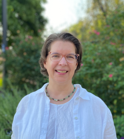 Becky Oskin, woman with glasses and a necklace wears a button down shirt. She smiles directly into the camera outside in front of bushes of flowers and trees.