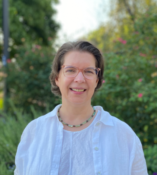 Becky Oskin, woman with glasses and a necklace wears a button down shirt. She smiles directly into the camera outside in front of bushes of flowers and trees. 