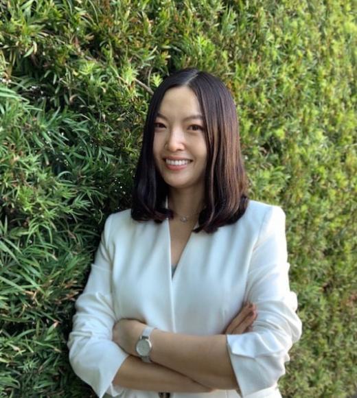 Woman smiling for a headshot in front of a leafy background while wearing a white blazer and a white watch