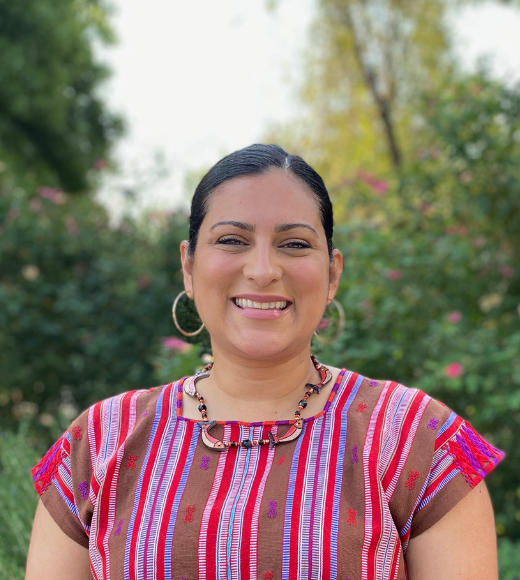 Gabriela Kovats Sanchez, woman with her hair back is wearing large earrings and a colorful necklace while smiling widely directly into the camera outside in front of bushes of flowers and trees.