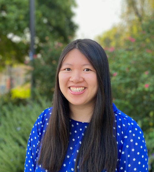 Karen Chan, woman wearing a polka dotted blouse smiles directly into the camera outside in front of bushes of flowers and trees. 