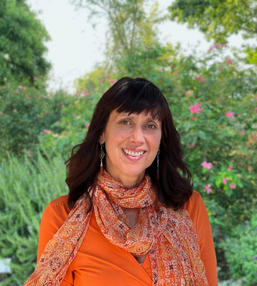 Moira Delgado, woman wearing earrings and a colorful scarf smiles widely directly into the camera outside in front of bushes of flowers and trees.