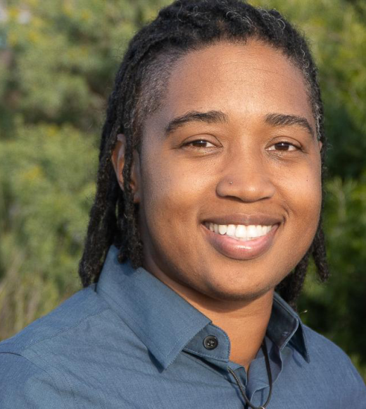 Close up of individual smiling outside. They are wearing a button down shirt.