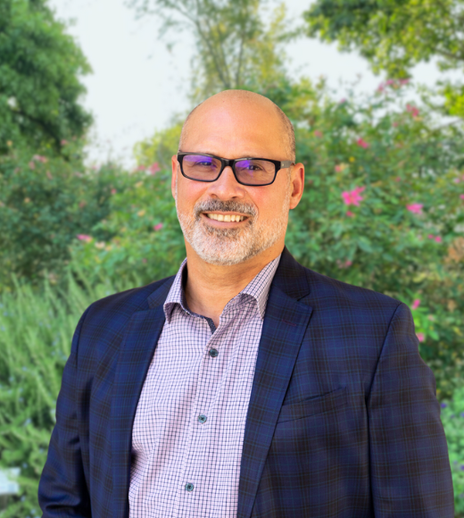 Michael Rios, man wearing tinted glasses and a plaid blazer and checkered dress shirt smiling directly into the camera outside in front of bushes of flowers and trees.