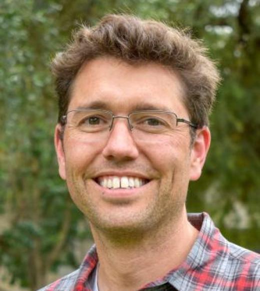 Man wearing glasses and a flannel plaid button up shirt smiles directly into the camera in front of a blurry nature background