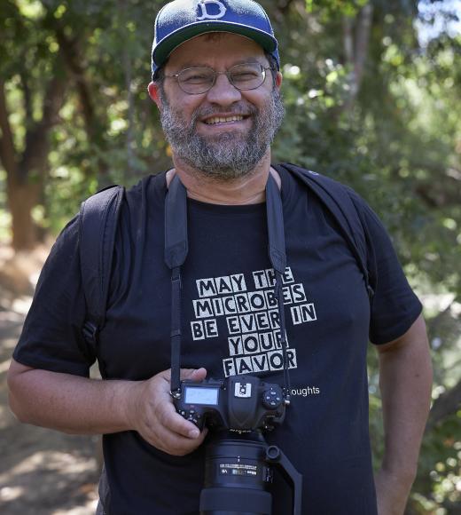 a man in a blue hat and glasses stands against a verdant green backdrop with a camera around his neck