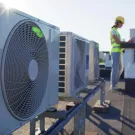 Oblique view of air conditioner units on a rooftop. A technician wearing a high visibility vest and hard hat is in the background. 