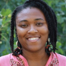 Close up headshot of a woman smiling outside. She is wearing a blouse with detailed beads on it.