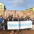 A group of about 40 people, most wearing coats and many wearing hats, stand together at a homebuilding site, behind a large white banner with the text "Build for Unity"